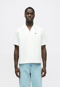 Krátky rukáv, biela košeľa s golierom s textúrovaným vzorom a zeleným logom Lacoste, doplnená svetlomodrými voľnými džínsami.