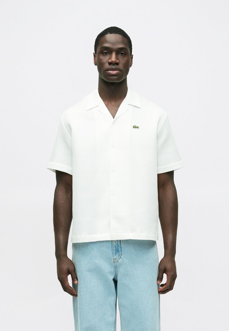 Krátky rukáv, biela košeľa s golierom s textúrovaným vzorom a zeleným logom Lacoste, doplnená svetlomodrými voľnými džínsami.