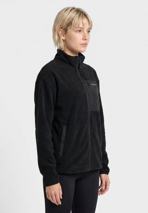 Ung kvinde med kort blondt hår iført en sort fleecejakke med lynlås og sorte bukser, stående sidelæns mod en ensfarvet baggrund.