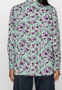 Pitkähihainen paitapusero, jossa on kukkakuosi purppuran, vihreän ja sinisen sävyissä valkoisella pohjalla; rento istuvuus ja nappikiinnitteiset kalvosimet.