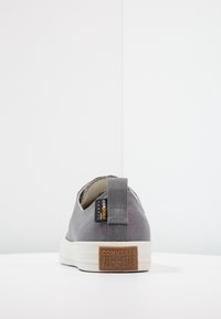 Šedá nízká teniska Converse All Star s bílou podešví, z pohledu zezadu, zobrazuje štítek z Cordura tkaniny a poutko na patě.