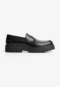 Zwarte leren instapper met stevige rubberen zool, moc-toe stiksels en decoratieve riemdetails op het gladde bovenwerk.