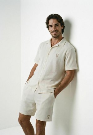 Un uomo si appoggia a una parete bianca indossando una camicia crema a maniche corte con bottoni e pantaloncini abbinati con un piccolo logo ricamato.
