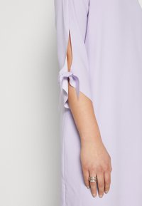 Robe lilas à manches longues et fentes sur les côtés, avec des nœuds aux poignets. Texture de tissu lisse et détails de conception épurés.