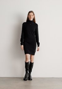 Vestido preto de gola alta feito de tecido de malha, design ajustado com mangas longas e corte até ao joelho, combinado com botas pretas até ao joelho.
