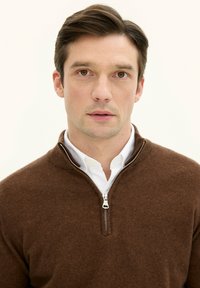 Brauner Reißverschluss-Pullover aus weichem Stoff, mit hohem Kragen und silbernem Reißverschluss. Über einem weißen Hemd mit Kragen getragen. Schlichte, strukturierte Oberfläche.