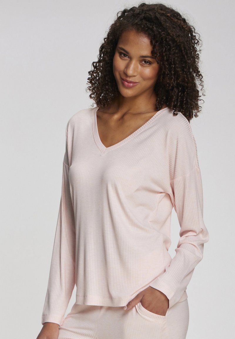 Woman with curly hair wearing a light pink ribbed long-sleeve top and matching pants, smiling with one hand in pocket.
