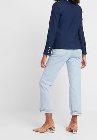 Tummansiniset istuvat bleiserit, joissa on kultaiset napit, yhdistettynä vaaleansinisiin, käännettyihin farkkuihin ja vaaleanruskeisiin kenkiin. Siistit viivat, sileä pinta ja jämäkkä muotoilu.