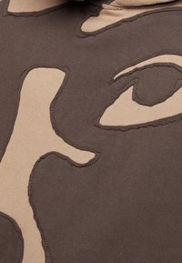 Primo piano di un tessuto marrone con forme astratte beige cucite per formare un volto parziale con dettagli degli occhi e del naso.