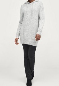 Robe à capuche en tricot gris avec une silhouette longue, cordons réglables et poignets côtelés, associée à un pantalon slim noir et des bottines.
