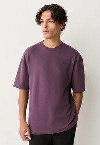 Lila T-Shirt mit kurzen Ärmeln und Rundhalsausschnitt, mit einem dezenten, geprägten Logo auf der Brust und einem lockeren Schnitt. Weiche Stoffstruktur.