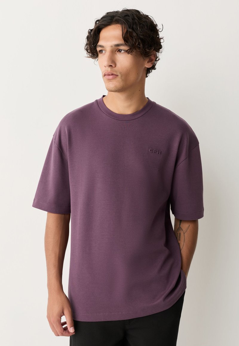 Lila T-Shirt mit kurzen Ärmeln und Rundhalsausschnitt, mit einem dezenten, geprägten Logo auf der Brust und einem lockeren Schnitt. Weiche Stoffstruktur.