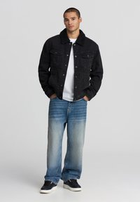 Giacca di jeans nera con colletto in peluche, abbinata a jeans a gamba larga sbiaditi di un blu chiaro e sneakers nere. Il modello sta in piedi con le mani nelle tasche.