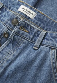 Vaqueros de mezclilla en azul con un acabado texturizado, que presentan dos botones metálicos, costuras amarillas y una etiqueta de marca en la cinturilla.