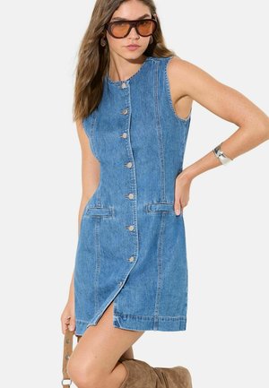 Kobieta w niebieskiej, jeansowej sukience na guziki bez rękawów, dużych brązowych okularach przeciwsłonecznych, srebrnej bransoletce, trzymająca beżowy pasek, pozująca z ręką na biodrze.