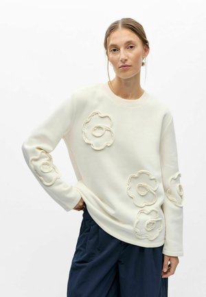 Jeune femme aux cheveux tirés en arrière portant un sweat-shirt blanc avec des motifs floraux en relief et un pantalon ample foncé sur un fond blanc uni.