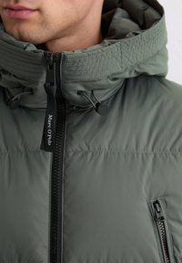 Roheline polsterdatud jakk kõrge kraega, lukuga sulgemine ja reguleeritavad kinnitusköied. Omab külgmist lukuga taskut ja brändimist lukukäepideme peal.