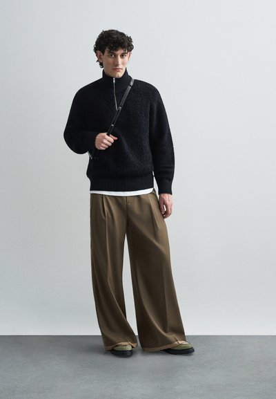 Schwarzer strukturierter Pullover mit hohem Kragen, kombiniert mit lockeren, khakifarbenen weit geschnittenen Hosen. Das Model hält einen schwarzen Riemen mit einer Tasche.