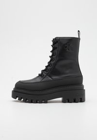 Černé kotníkové boty s kulatou špičkou, texturovaným koženým svrškem, vyvýšenou platformou a výrazným vzorkem podrážky, se šněrováním a logem.