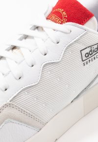 White Adidas Supercourt sneaker with red tongue, white laces, and textured fabric side panel on a white background.