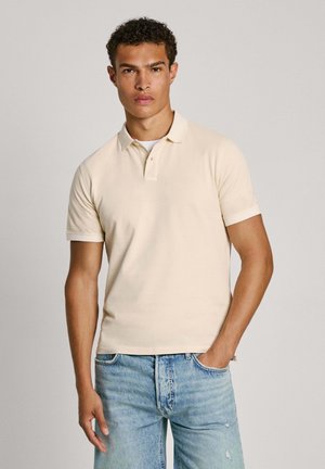 Camisa polo beige hecha de algodón texturizado, con un cuello clásico, mangas cortas y un dobladillo recto. Se lleva con jeans desgastados de color azul claro.