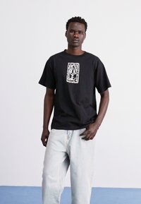 Camiseta de algodón negra con un diseño gráfico blanco que presenta una forma puntiaguda y un borde decorativo. Llevada con jeans de mezclilla azul claro.