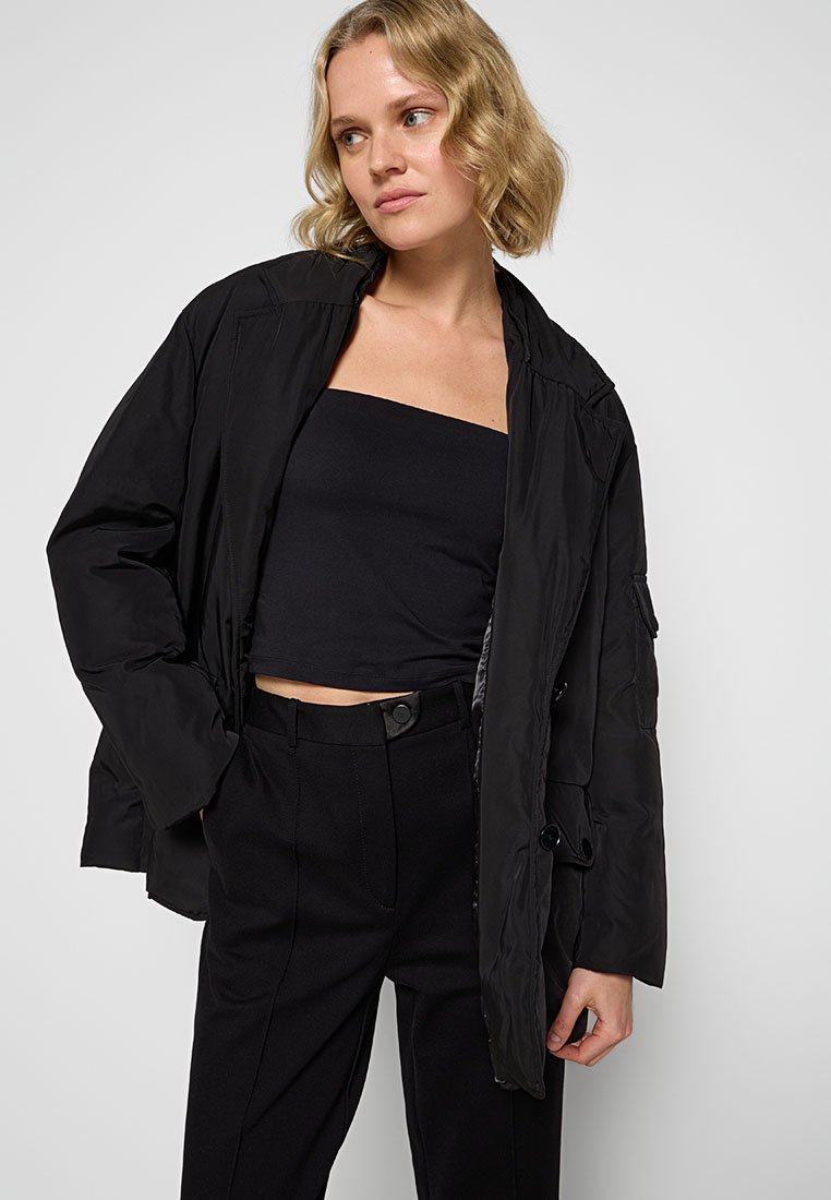 Schwarze Pufferjacke mit Reißverschluss- und Knopfverschluss, ausgestattet mit Seitentaschen und einer lässigen Passform, kombiniert mit einem figurbetonten schwarzen Oberteil und einer Hose.