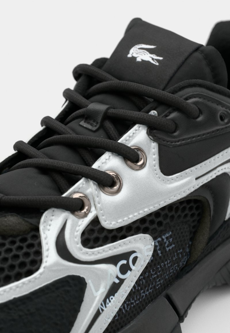 Zapatilla deportiva negra y plateada con parte superior de malla texturizada, diseño de cordones, ojales de metal y detalle de logo en relieve en la lengua.