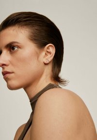 Pendientes de gota plateados con una cadena y un colgante de barra recta. El modelo tiene el cabello corto y oscuro, y lleva una blusa sin mangas en color topo.