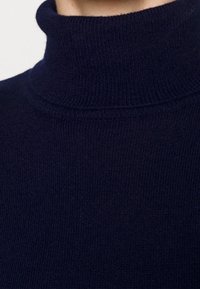 Tmavě modrý rolák vyrobený z texturované pleteniny, s ribbovaným límcem a bezšvým designem, který ukazuje rovnoměrné stehy.