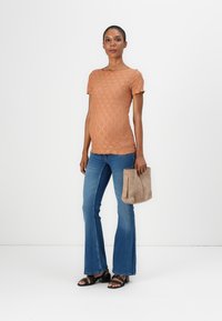 Lys orange teksturert t-skjorte med korte ermer, kombinert med utsvingte blå jeans og svarte stroppesandaler, holder en beige håndveske.