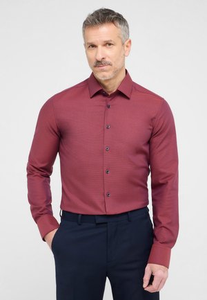 Bordeauxfarbenes Langarmhemd aus strukturiertem Stoff, mit Button-Down-Kragen und blauen Knöpfen. Kombiniert mit einer schmal geschnittenen blauen Hose.
