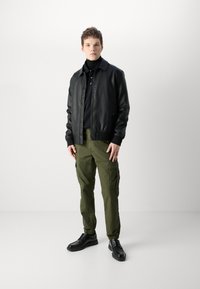 Casaco bomber de couro preto sobre um suéter preto de gola alta, combinado com calças cargo verde-oliva e sapatos pretos de atacadores. Design simples, com bolsos funcionais.