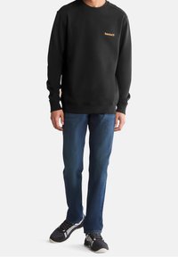 Felpa nera con polsini e orlo a costine, caratterizzata da un piccolo logo arancione sul petto. Abbinata a jeans blu scuro e sneakers nere.