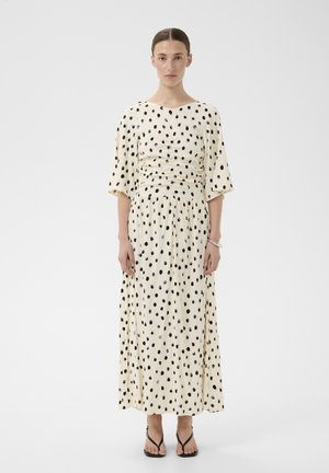 Femme debout dans une robe midi couleur crème à pois noirs, manches mi-longues et sandales noires ouvertes à l'avant sur fond blanc.