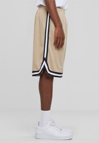 Shorts de sport beiges avec des bandes latérales noires et blanches. Conçus en tissu mesh, avec une ceinture élastique et une coupe ample.