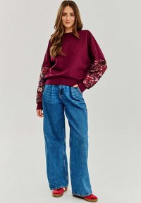 Burgundowa bluza z kwiecistymi rękawami, połączona z szerokimi niebieskimi dżinsami i czerwonym obuwiem. Miękka tkanina w luźnym kroju.