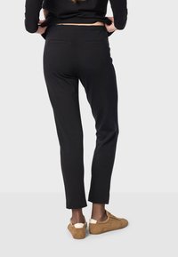 Pantalon noir avec une coupe ajustée, comprenant deux poches arrière et une texture lisse. Associé à des chaussures beiges pour un contraste.