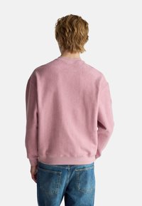 Różowa bluza polarowa z okrągłym dekoltem i ściągaczami na rękawach, zestawiona z jasnoniebieskimi dżinsami. Materiał wydaje się miękki i teksturowany.