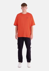 Oranges Übergrößen-T-Shirt mit kurzen Ärmeln, schwarze Cargo-Hosen mit Taschen und weiße Sneakers. Einfaches Design mit minimaler Detailgestaltung.