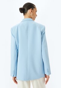 Lichtblauwe oversized blazer met gestructureerde schouders, scherpe revers en vier knoppen op de mouwen. Soepele stof en getailleerd silhouet.