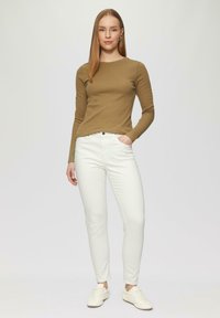 Bruine geribbelde langslavenshirt gecombineerd met witte aansluitende jeans en witte sneakers, staand tegen een lichtgrijze achtergrond.