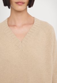 Suéter de punto beige con escote en V, textura acanalada y costuras de hombro caídas, que destacan su apariencia suave y cálida.