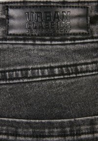 Tela vaquera negra con superficie texturizada, que presenta una etiqueta de cuero estampada con "URBAN CLASSICS DENIM FACTORY" en la cintura.