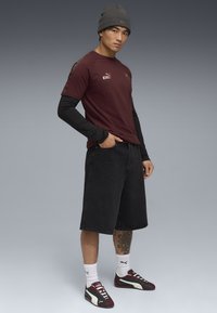 Camisa de manga corta marrón con logo, llevada sobre una camisa de manga larga negra. Pantalones cortos de mezclilla negros, zapatillas burdeos y verdes, gorra de punto gris.
