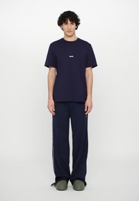 T-shirt in cotone blu navy con maniche corte e logo bianco, abbinato a pantaloni della tuta blu navy con strisce laterali e zoccoli verdi.