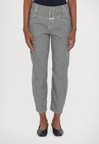Pantalones a rayas con rayas verticales en blanco y negro, pierna ajustada, bolsillos frontales, cremallera y zapatos planos negros. Textura de tela suave.