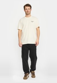 Beige katoenen t-shirt met korte mouwen, voorzien van een kleine grafische print op de borst, gecombineerd met zwarte jeans en een paar bruine sneakers.