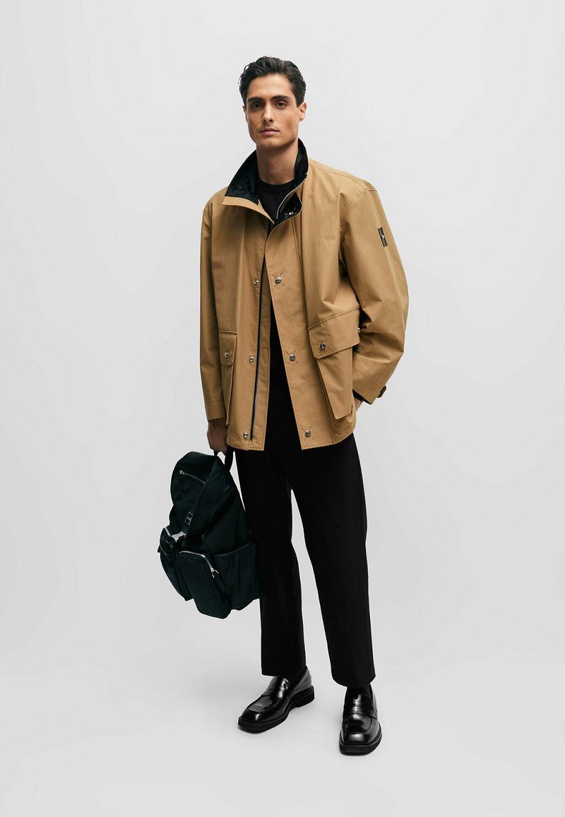 Beige wasserdichte Jacke mit schwarzem Kragen, ausgestattet mit Druckknöpfen und zwei Fronttaschen, getragen mit schwarzen Hosen und einem grünen Rucksack.