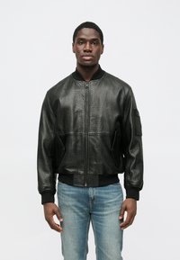 Mustast nahast bomber-tüüpi jakk tekstuurse viimistlusega, luku sulgemisega, soonikkäiste ja küljetaskutega. Ühildatud hele siniste teksadega.